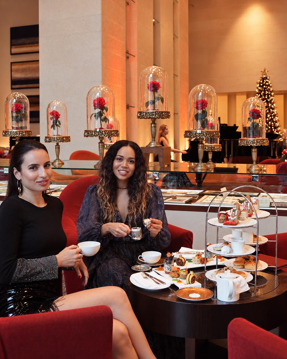 Rosi Ross and Vasimav seated together at Raffles Dubai during a Christmas afternoon tea, surrounded by tiered pastry stands, teacups, festive desserts, and elegant red lounge seating.