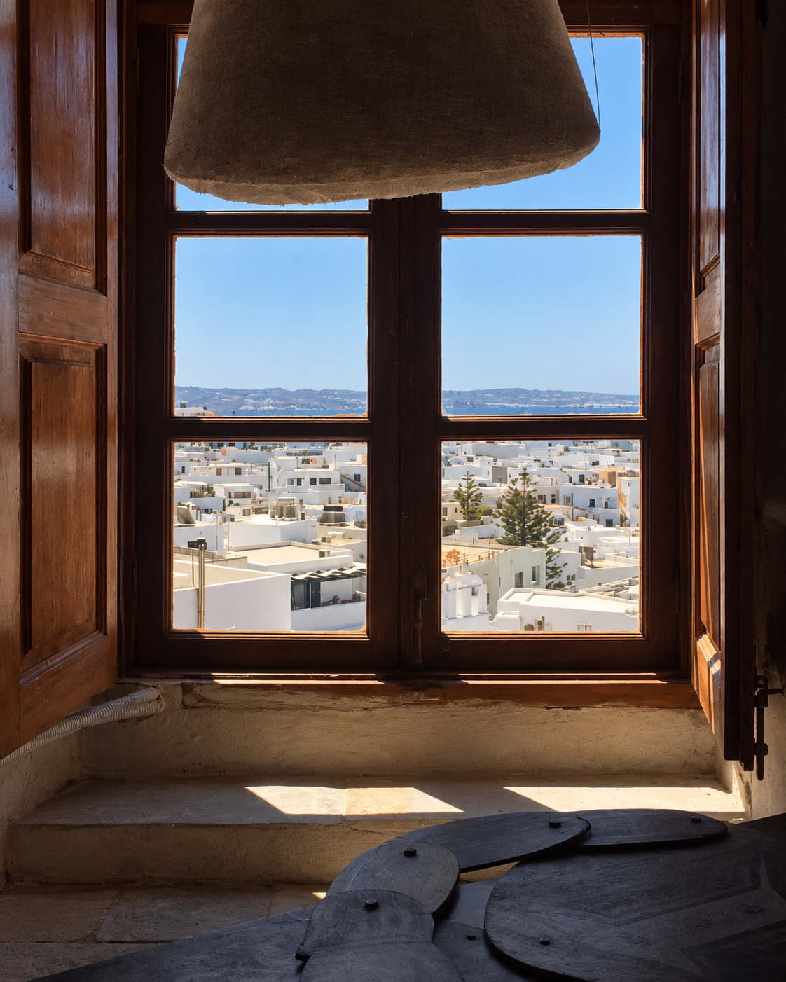 View through a traditional wooden-framed window at the Archaeological Museum of Naxos, overlooking whitewashed buildings and the town of Chora beneath a clear Mediterranean sky.