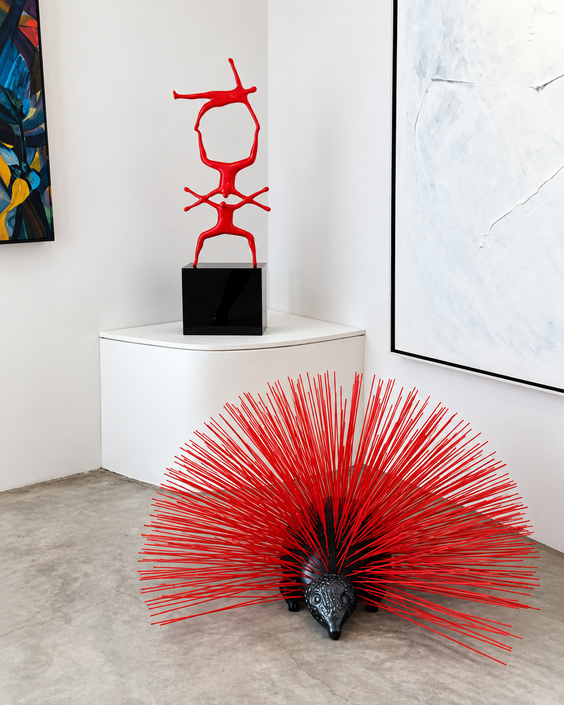 Contemporary art gallery in Mykonos displaying a vibrant red spiky sculptural installation in the foreground and an abstract red sculpture on a white pedestal against minimalist gallery walls.