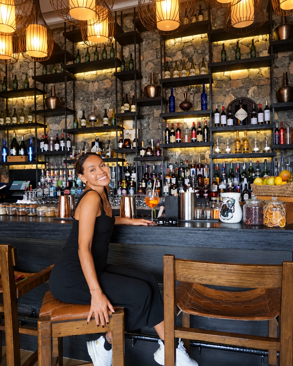 Alila Jabal Akhdar Rosi Ross Smiling at the Warm Rustic Bar Rosi Ross seated at the bar at Alila Jabal Akhdar, Oman, surrounded by stone walls, illuminated liquor shelves and warm pendant lighting.