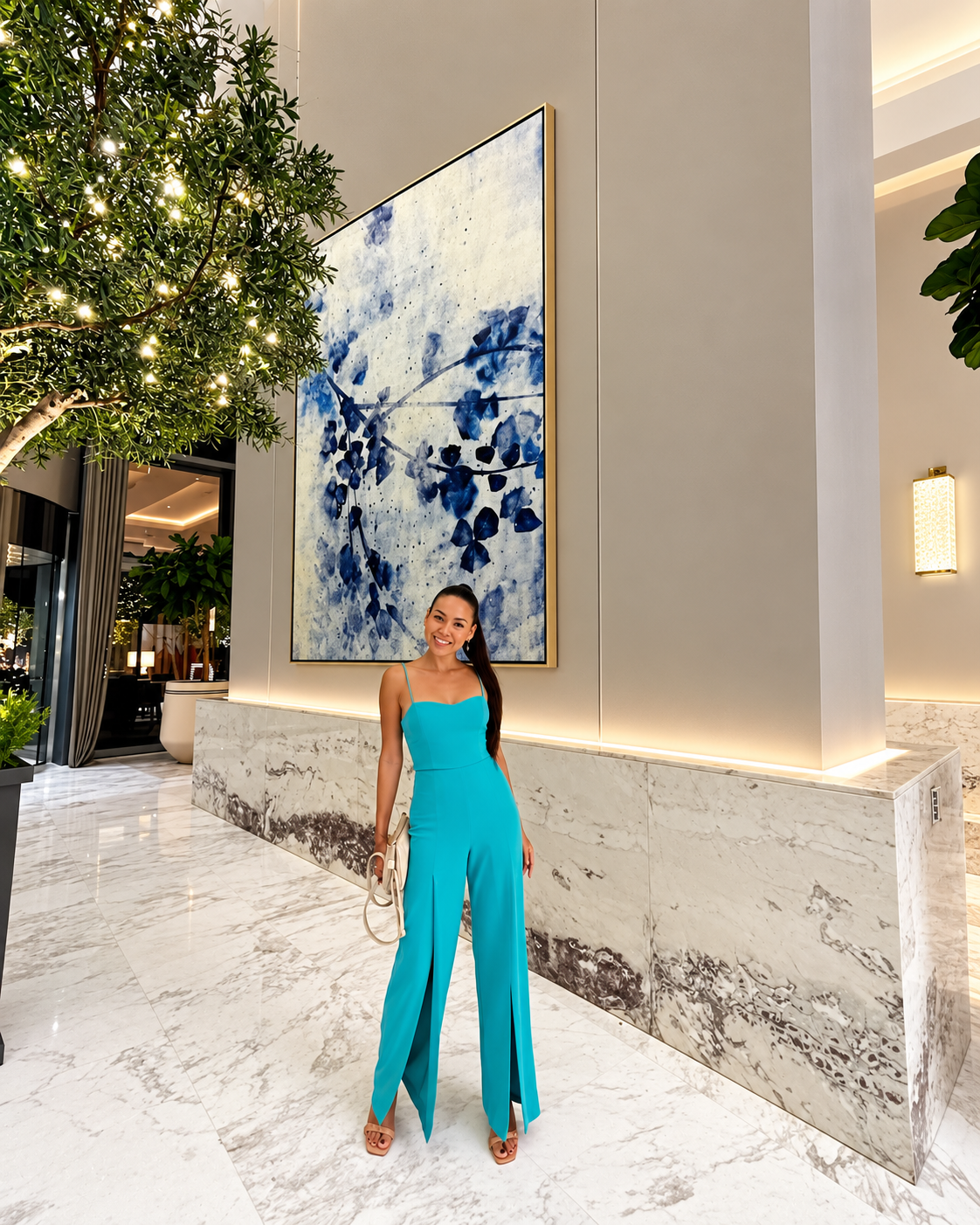 Rosi Ross in Revolve teal jumpsuit posing in elegant lobby with marble floors and large abstract blue art.