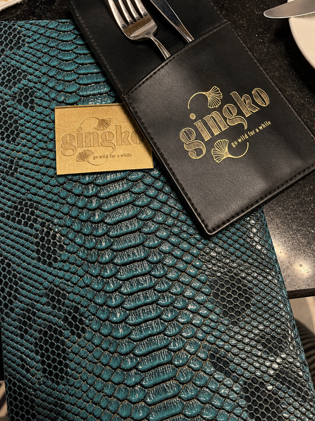 Gingko restaurant menu and black leather cover with gold text on a teal snake skin patterned surface.