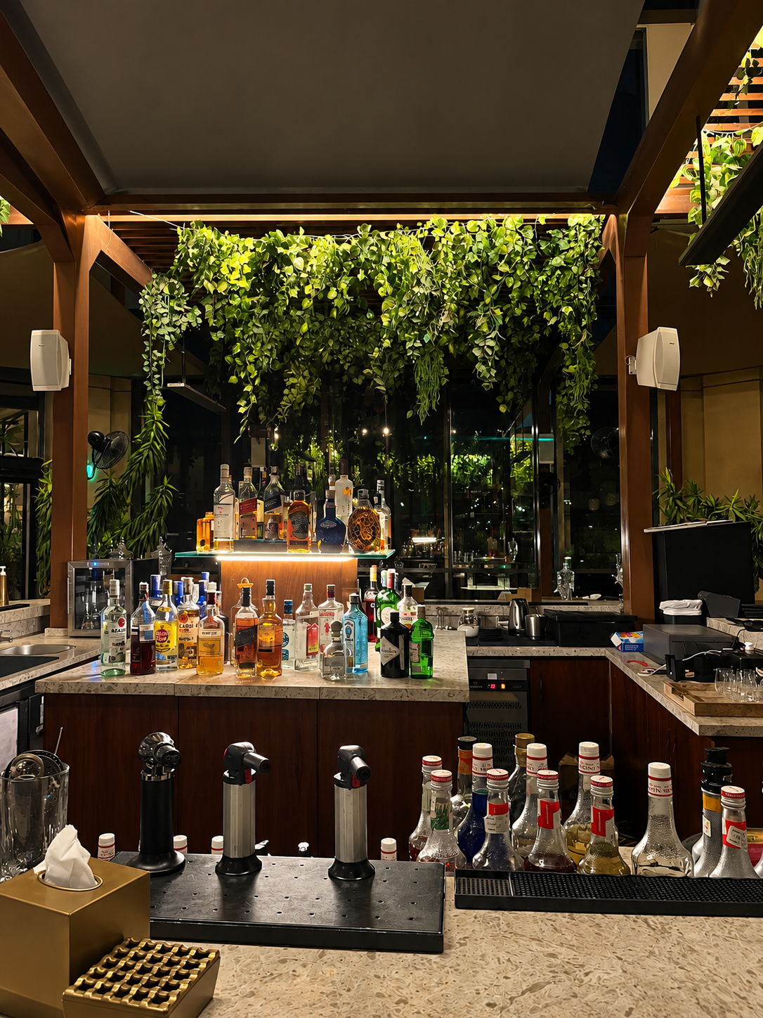 Upscale bar with assorted liquor bottles, hanging greenery, and sleek countertops.