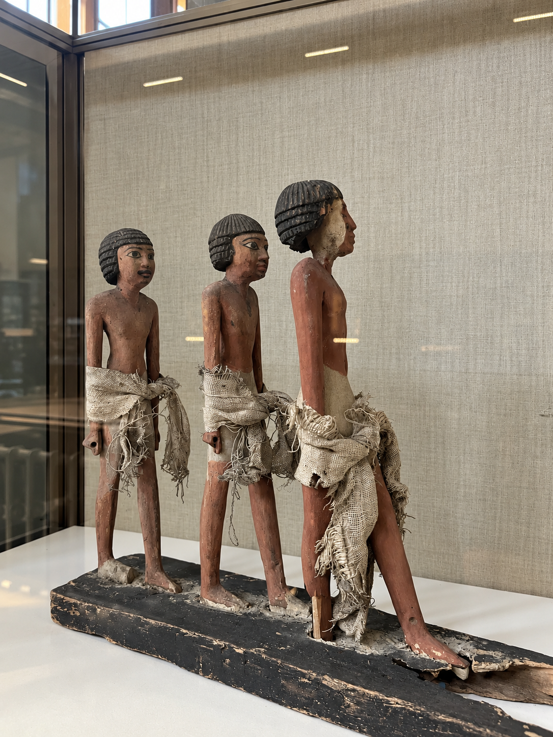 Ancient Egyptian Wooden Figurines at the National Museum of Egyptian Civilization in Cairo Three ancient Egyptian wooden figurines displayed in a glass case, depicting standing male figures with linen garments at the National Museum of Egyptian Civilization in Cairo