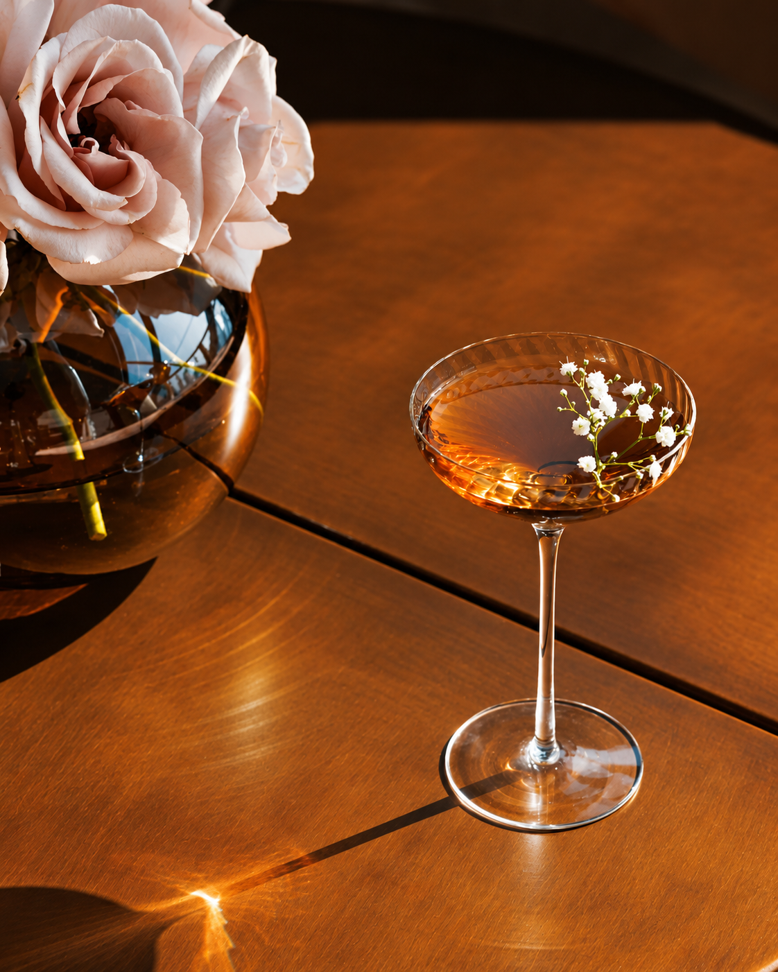 Elegant cocktail in a coupe glass with baby's breath garnish beside pink roses on a brown table.