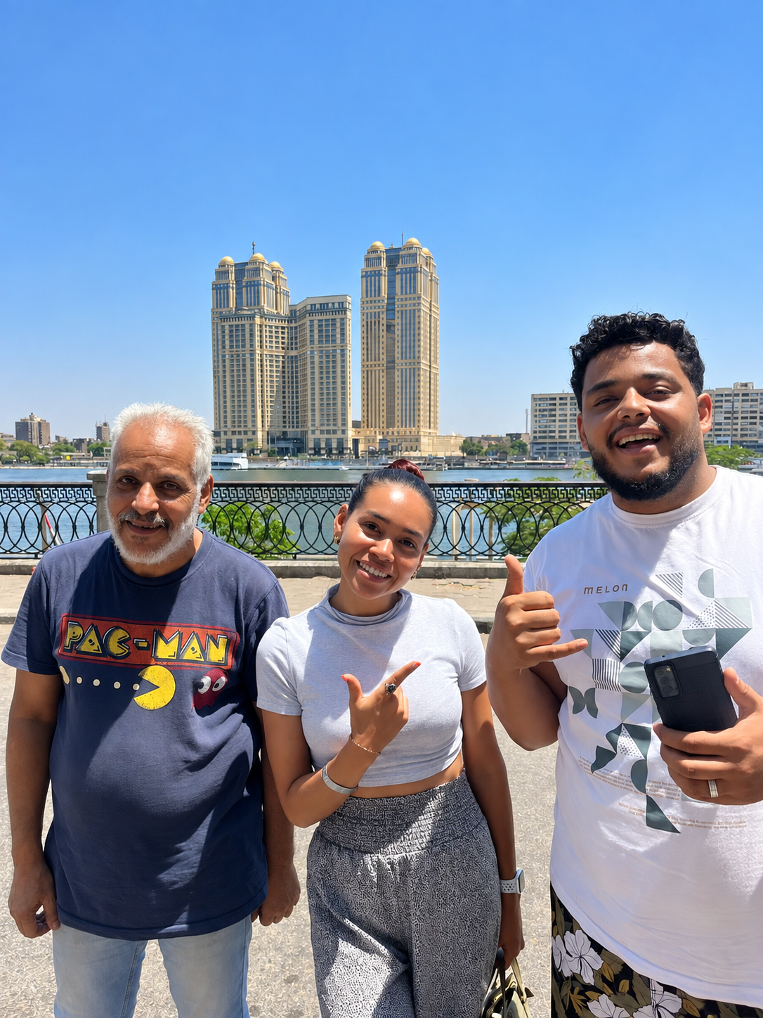 Rosi Ross And Ahmed Family Driver In Cairo Ahmed, his father, and Rosi Ross pose for a photo with a river and luxury hotel buildings in the background under a clear blue sky.