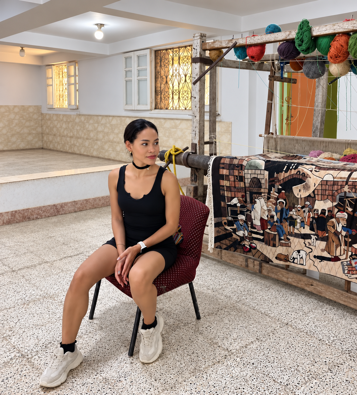 Rosi Ross sits thoughtfully in a carpet weaving workshop at the Carpet School in Saqqara Rosi Ross in black dress and sneakers sitting in a traditional carpet weaving workshop with colorful yarn and a loom at the Carpet School in Saqqara