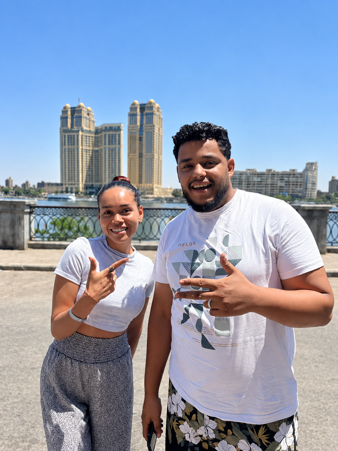 Rosi Ross and Ahmed At Drop Off In Cairo Rosi Ross and Ahmed smiling, posing in front of modern high-rise buildings and a body of water under a clear blue sky.