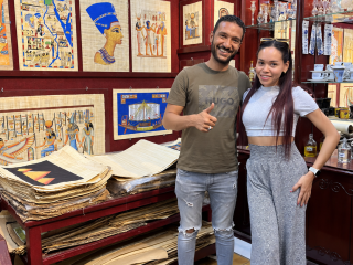 Rosi Ross and Mohammad smiling in a papyrus shop, surrounded by Egyptian artworks and papyrus sheets.