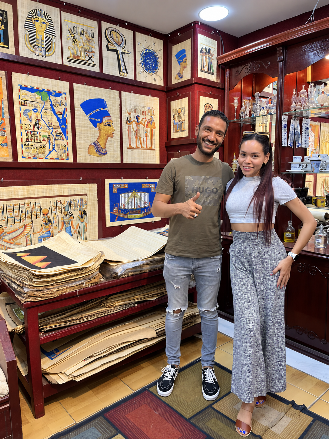 Rosi Ross and Mohammad smiling in a papyrus shop, surrounded by Egyptian artworks and papyrus sheets.