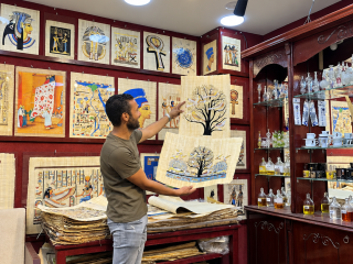 Mohammas in store holding a papyrus painting of a tree, surrounded by ancient Egyptian artwork.