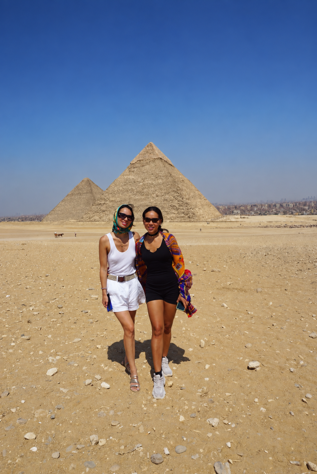 Rosi Ross and Vasimav at the Saqqara Pyramids Rosi Ross and Vasimav pose in front of the Great Pyramids of Giza under a clear blue sky, one wearing white shorts and a tank top with a headscarf, the other in a black dress with a colorful kimono.