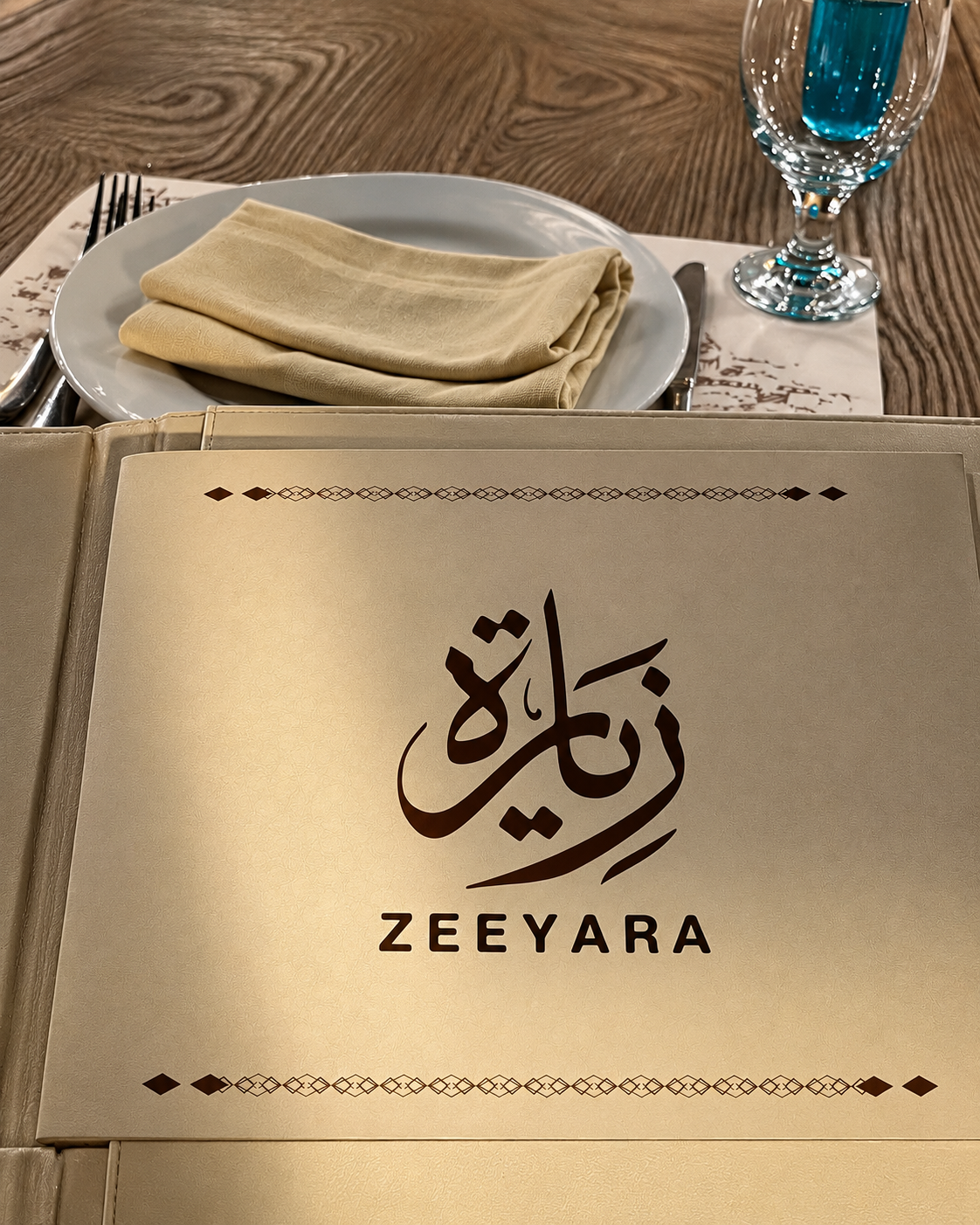 Zeeyara restaurant menu on a wooden table with a plate, utensils, and a glass of blue liquid.
