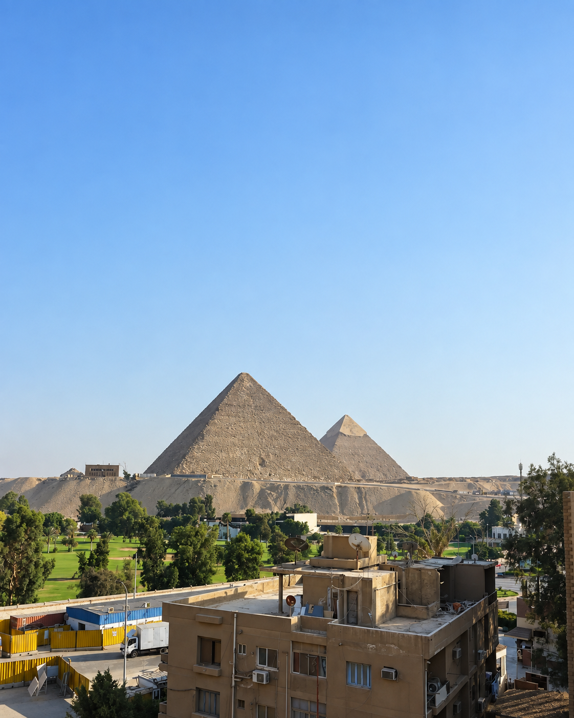 Panoramic view of the Giza Pyramids and urban landscape with buildings, trees, and desert under a clear sky.
