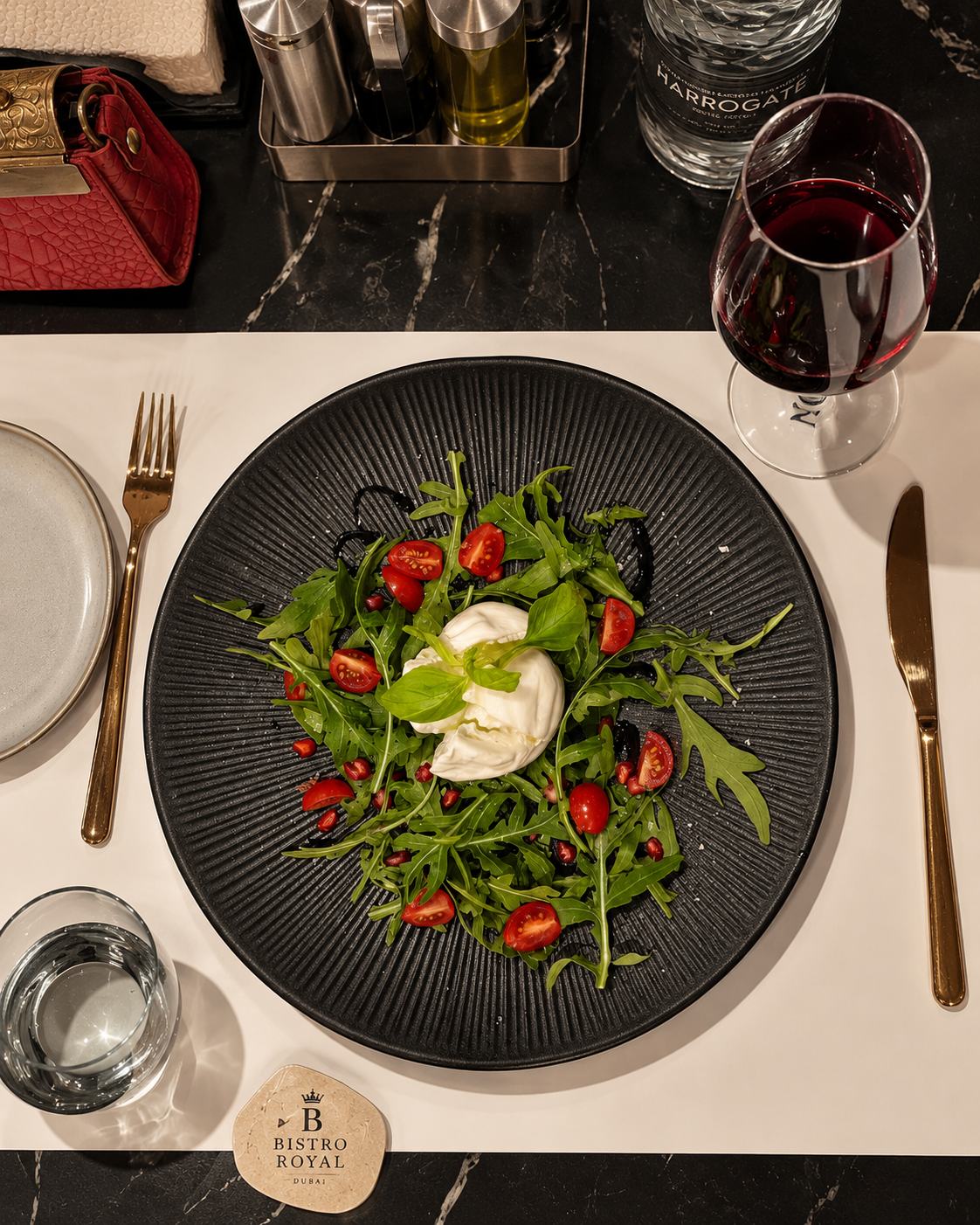 Burrata Salad with Arugula and Cherry Tomatoes at Bistro Royal Design District Arugula salad with burrata cheese, cherry tomatoes, and pomegranate seeds on a black plate, accompanied by a glass of red wine at Bistro Royal Design District
