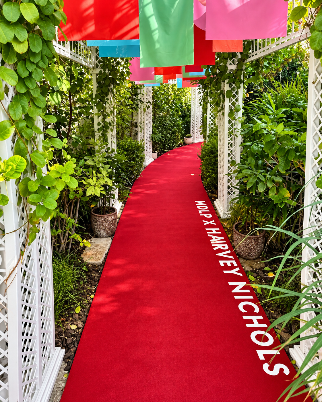 Red carpet pathway through white lattice garden structure with lush green vines and colorful overhead panels at MDLP x Harvey Nichols Event