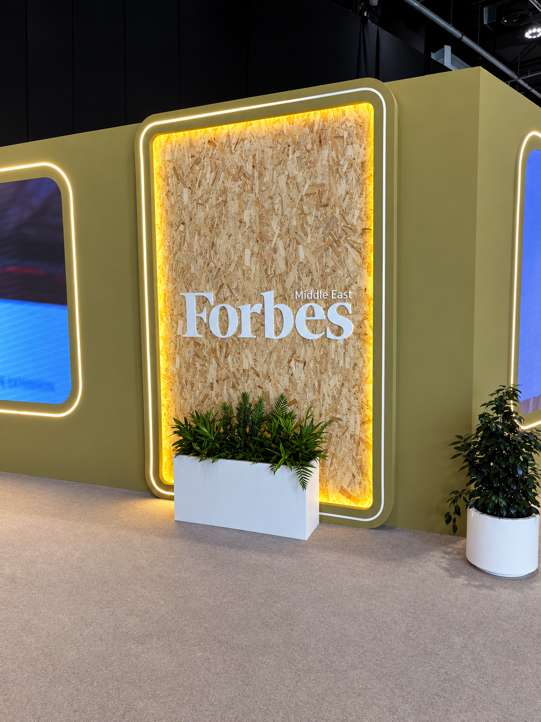 RosiRoss Forbes Sustainability Summit AD Ambiance A Forbes Middle East logo sign with a wood chip texture background, illuminated by a warm yellow light, beside a planter.