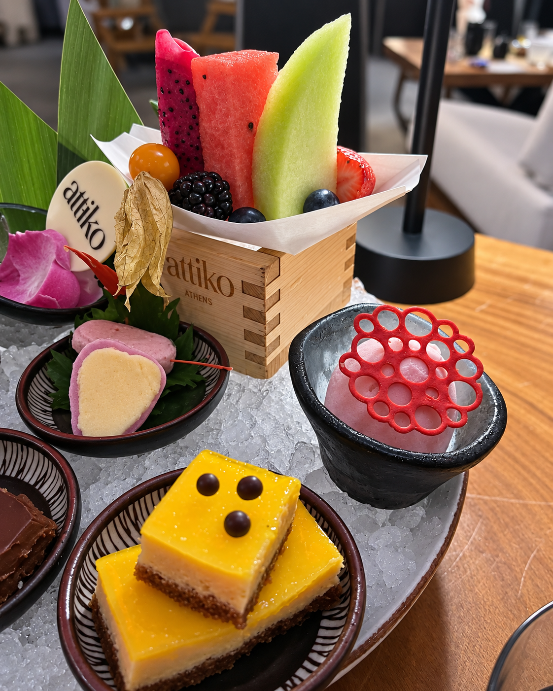 Fine Dining Dessert Platter with Fresh Fruit at ATTIKO Dubai Fine dining dessert platter featuring fresh watermelon and pear slices in a wooden box, colorful gelatin domes, and yellow glazed cakes on purple plates at ATTIKO Dubai