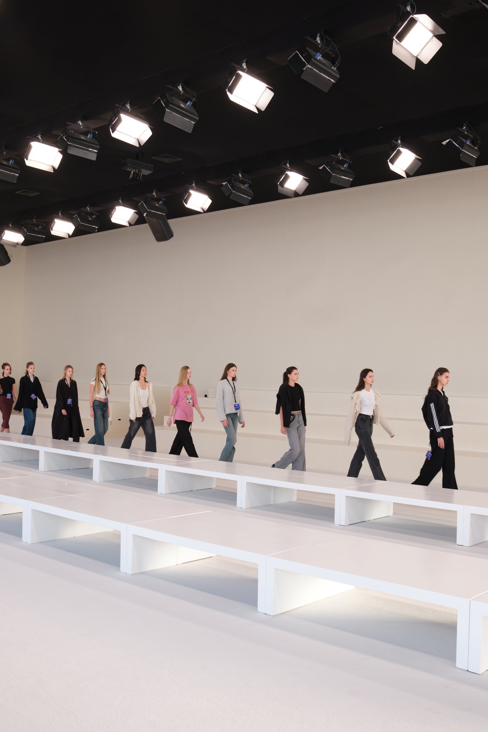 Models walking in single file line on white runway with professional overhead stage lights and white bench seating
