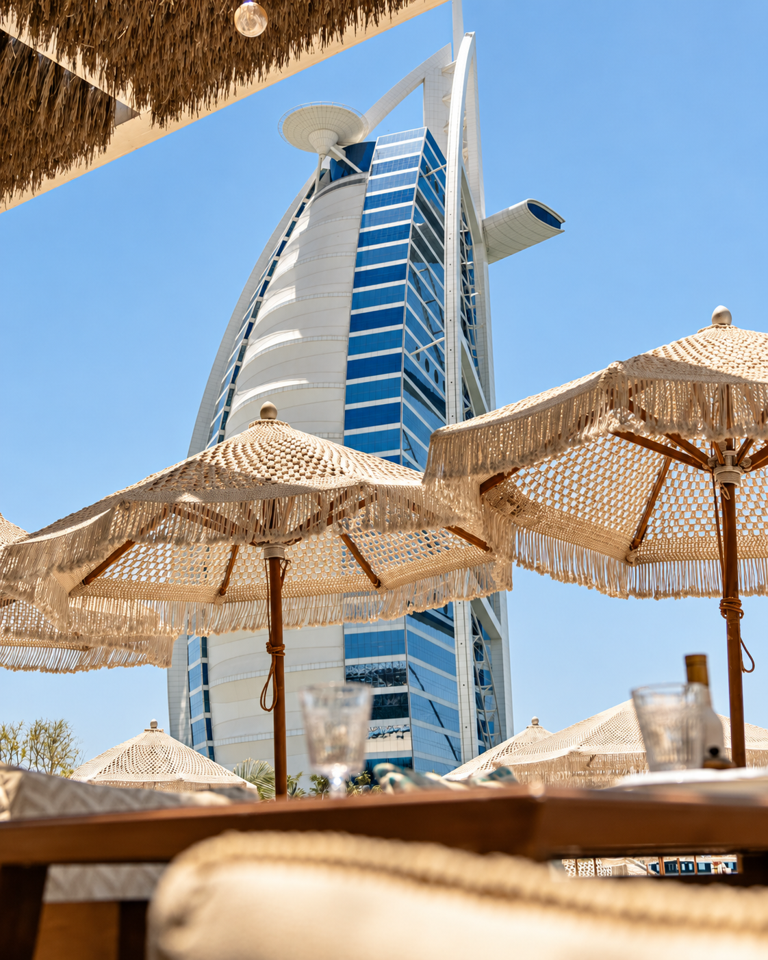 Burj Al Arab view from the Nuska beach with beige fringed umbrellas overlooking a white sail-shaped resort tower under clear blue sky