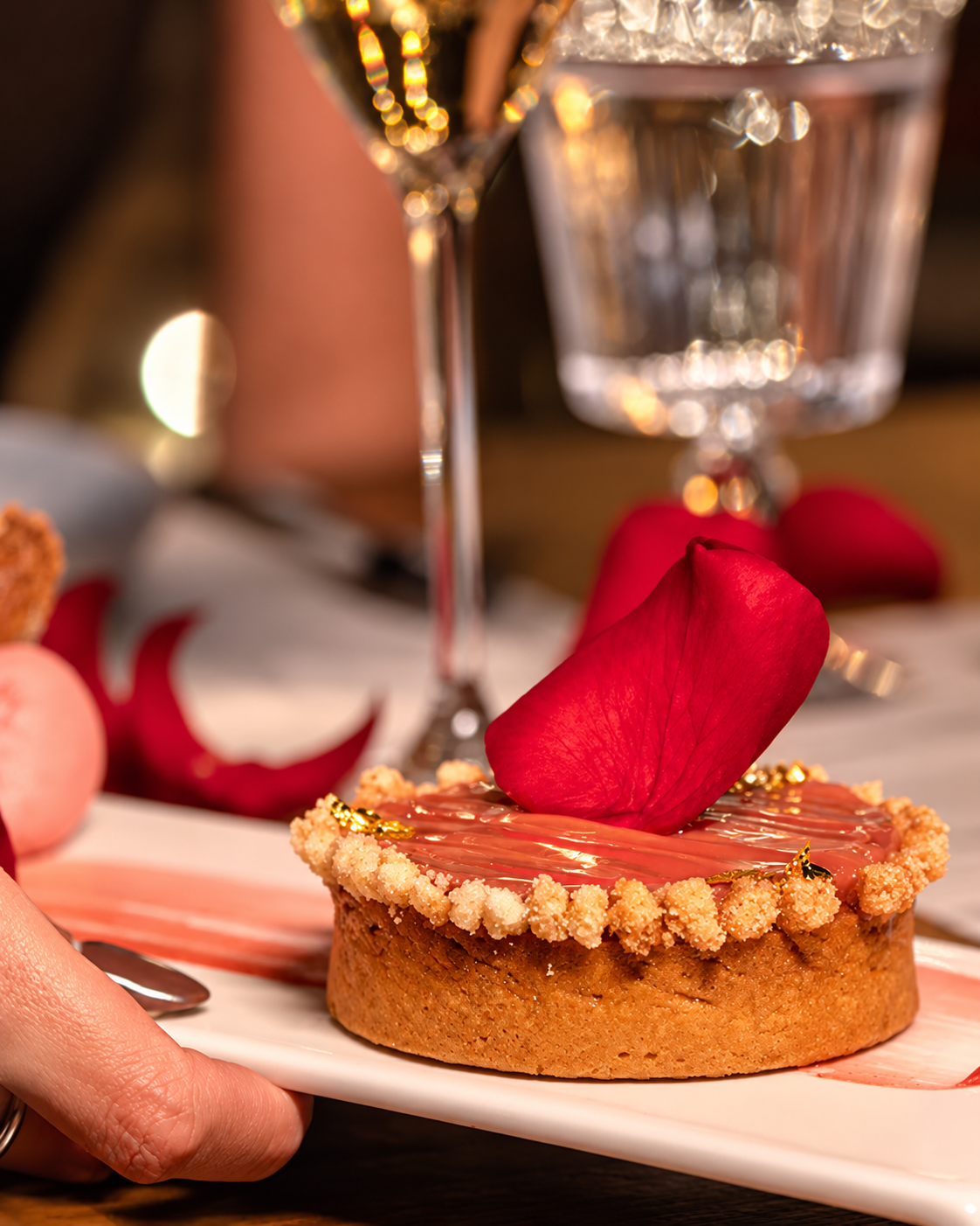Nuska Elegant Dessert with a red rose petal and gold leaf, served on a white plate with champagne and ambient lighting.
