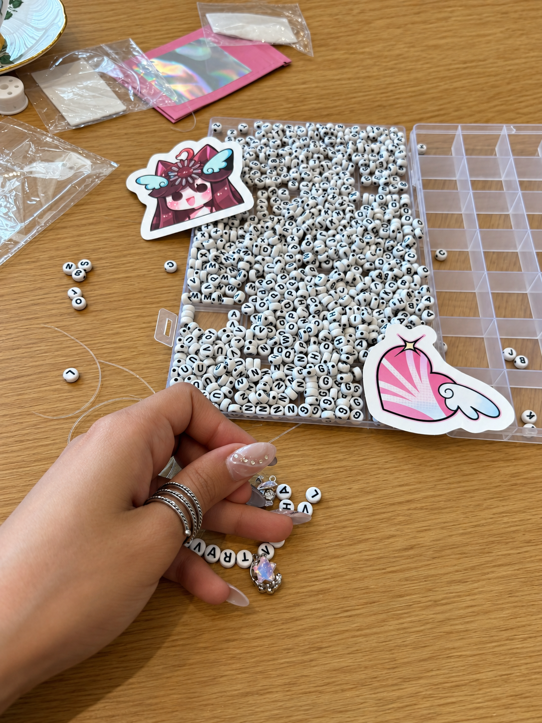 Rosi Ross close-up of hands creating a charm bracelet with letter beads and iridescent charms, alongside a bead organizer on a wooden table.