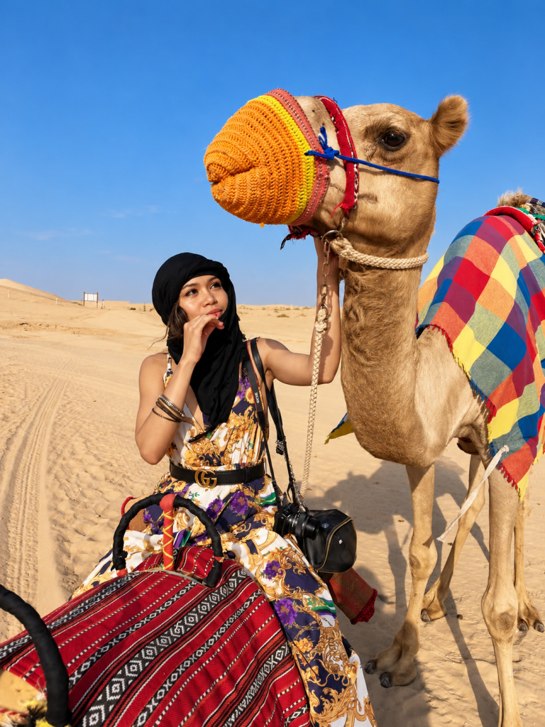 Rosi Ross beside a camel wearing a colorful woven muzzle and saddle blanket at Al Marmoom Desert Dubai, dressed in a printed outfit and black headscarf.